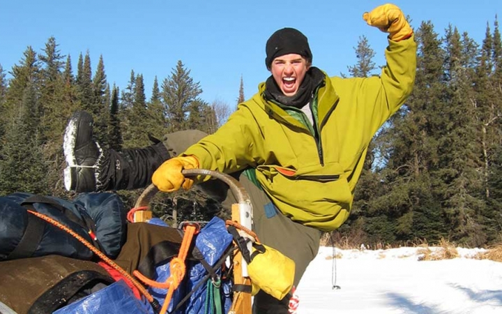 A person flexes their muscles while standing at the back of a dog sled